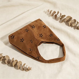 May include: A brown suede handbag with a single handle. The bag features a floral cut-out design across the front. The bag is set against a neutral background with dried foliage.
