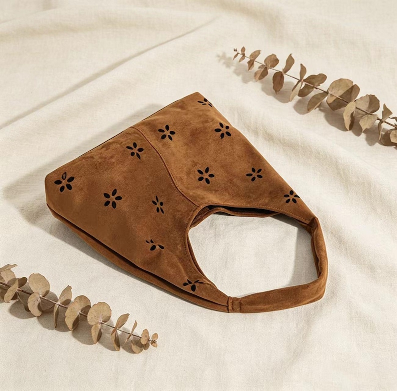 May include: A brown suede handbag with a single handle. The bag features a floral cut-out design across the front. The bag is set against a neutral background with dried foliage.
