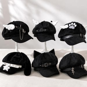 Puede incluir: Seis gorras negras con varios adornos. Algunas gorras presentan alas blancas, una huella de pata o orejas de gato. Otras tienen dijes plateados y una correa blanca. Cada gorra tiene una visera negra y una correa decorativa.