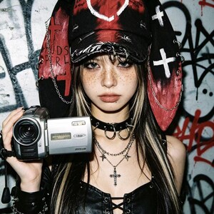May include: A person wearing a black and red hat with a heart design and cross accents, holding a silver camcorder. The person is also wearing a black choker, layered necklaces with crosses and stars, and a black leather top.