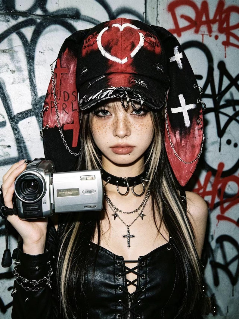 May include: A person wearing a black and red hat with a heart design and cross accents, holding a silver camcorder. The person is also wearing a black choker, layered necklaces with crosses and stars, and a black leather top.