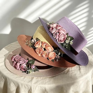 Puede incluir: Cuatro sombreros de paja apilados en tonos rosa, naranja, amarillo y morado. Cada sombrero está adornado con un racimo de rosas rosas y vegetación. Los sombreros tienen alas anchas y están dispuestos de forma atractiva.