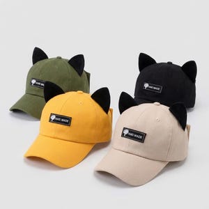 Puede incluir: Cuatro gorras de béisbol en verde oliva, amarillo, negro y beige, cada una con orejas de gato negras y un parche "HAND MADE". Las gorras se muestran sobre un fondo blanco.