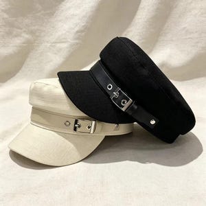 Puede incluir: Dos gorras de estilo marinero, una negra y otra color blanco roto, ambas con un acabado de tela texturizada. Cada gorra incluye una tira decorativa en la parte delantera con una hebilla de metal plateado y detalles de ojales metálicos.