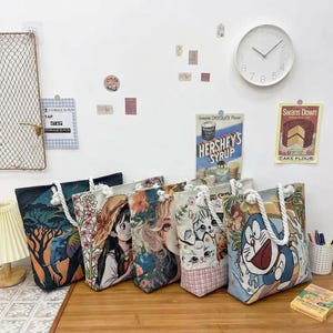 Puede incluir: Una colección de bolsos tote con varios diseños coloridos, incluyendo ilustraciones de personajes de anime, animales y patrones florales. Cada bolso tiene un asa de cuerda blanca. Los bolsos se exhiben sobre una superficie de madera.