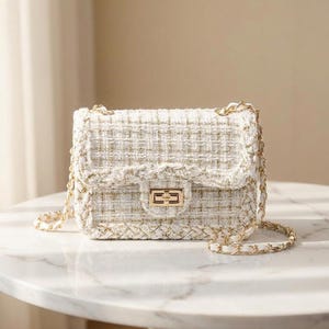 Puede incluir: Pequeño bolso de mano de tweed blanco con detalles dorados. El bolso tiene una correa de cadena dorada y un cierre dorado. La tela de tweed tiene una textura tejida con hilos dorados. El bolso es rectangular con solapa.