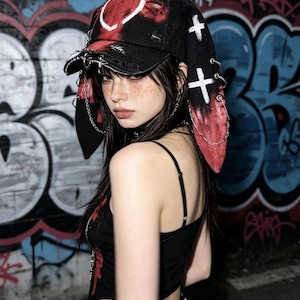 May include: Black baseball cap with a red and white heart design, distressed details, and long, red-tipped ear flaps. The cap features white cross accents and silver chain embellishments, creating a unique, edgy aesthetic.