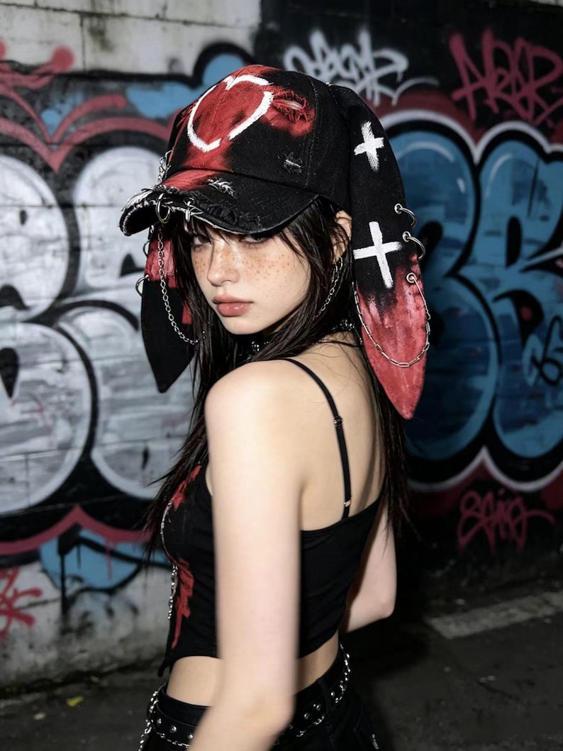 May include: Black baseball cap with a red and white heart design, distressed details, and long, red-tipped ear flaps. The cap features white cross accents and silver chain embellishments, creating a unique, edgy aesthetic.