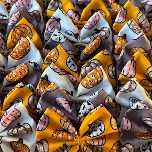 May include: A collection of fabric hair bows featuring a repeating pattern of pumpkins in shades of orange, white, and muted purple. Each bow is gathered in the centre, showcasing a variety of pumpkin illustrations printed on the textile surface.