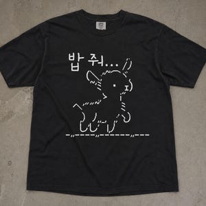 May include: A black short-sleeve t-shirt featuring a white ASCII art graphic of a small animal standing on a dashed line. Above the animal, the Korean text 밥 줘... is printed in white. The shirt has a crew neckline and a standard fit.