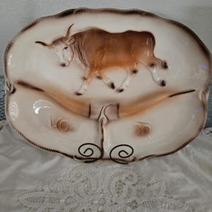 May include: Oval-shaped ceramic serving platter with a raised relief of a brown bull. The platter has a cream-colored background with brown accents and the letters "LO" and "BO". The platter is supported by two black metal scrollwork feet.