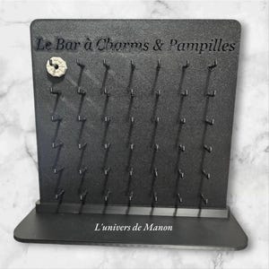 Peut inclure: Présentoir noir avec de nombreux crochets pour bijoux ou accessoires. Le texte "Le Bar à Charms & Pampilles" et "L'univers de Manon" sont imprimés sur le présentoir. Un charm décoratif est fixé en haut à gauche.