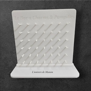 Peut inclure: Présentoir blanc avec de nombreux petits crochets, conçu pour accrocher des breloques ou des articles similaires. Le texte "Le Bar à Charms & Pampilles" est en relief en haut, et "L'univers de Manon" en bas.
