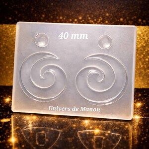 Puede incluir: Un molde de silicona transparente y rectangular con dos diseños en espiral y dos pequeños círculos. El texto "40 mm" está en la parte superior y "Univers de Manon" en la inferior. El fondo es un degradado de oro y negro.