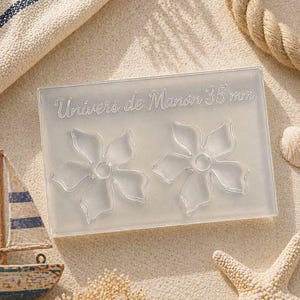 Peut inclure: Un moule en silicone transparent rectangulaire avec deux cavités en forme de fleur. Le texte "Univers de Manon 35 mm" est en relief sur le dessus. Le moule est posé sur une surface sablonneuse avec des décorations nautiques.