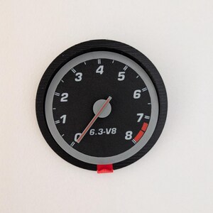 Peut inclure: Un indicateur noir et gris avec une aiguille rouge, affichant les chiffres de 1 à 8. L'indicateur indique "6.3-V8" et comporte un indicateur rouge au chiffre 7. L'indicateur est monté sur une surface blanche.