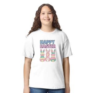 Könnte beinhalten: Weißes T-Shirt mit dem Text "HAPPY EASTER" in Blau, Rosa und Lila. Darunter befinden sich drei bunte Hasenillustrationen mit Zickzack-, Karomustern und Streifen. Das T-Shirt wird von einer Person getragen.