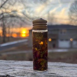 Op de afbeelding: Een kleine, heldere glazen fles met een goudkleurige dop, gevuld met een vloeistof en bloemblaadjes. De fles staat op een houten oppervlak, met een vage achtergrond van een zonsondergang en een huis.