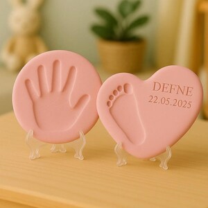 May include: Pink keepsake handprint and footprint molds on clear stands. One mold is round with a handprint, the other is heart-shaped with a footprint and the name "DEFNE" and the date "22.05.2025".