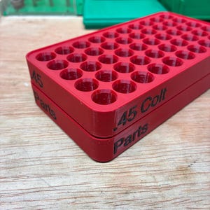 Pode incluir: Uma bandeja de armazenamento retangular de pl&aacute;stico vermelho com uma grade de orif&iacute;cios circulares para organizar muni&ccedil;&otilde;es. A lateral da bandeja apresenta texto impresso em preto que diz .45 Colt e Parts. Design estruturado e em camadas.