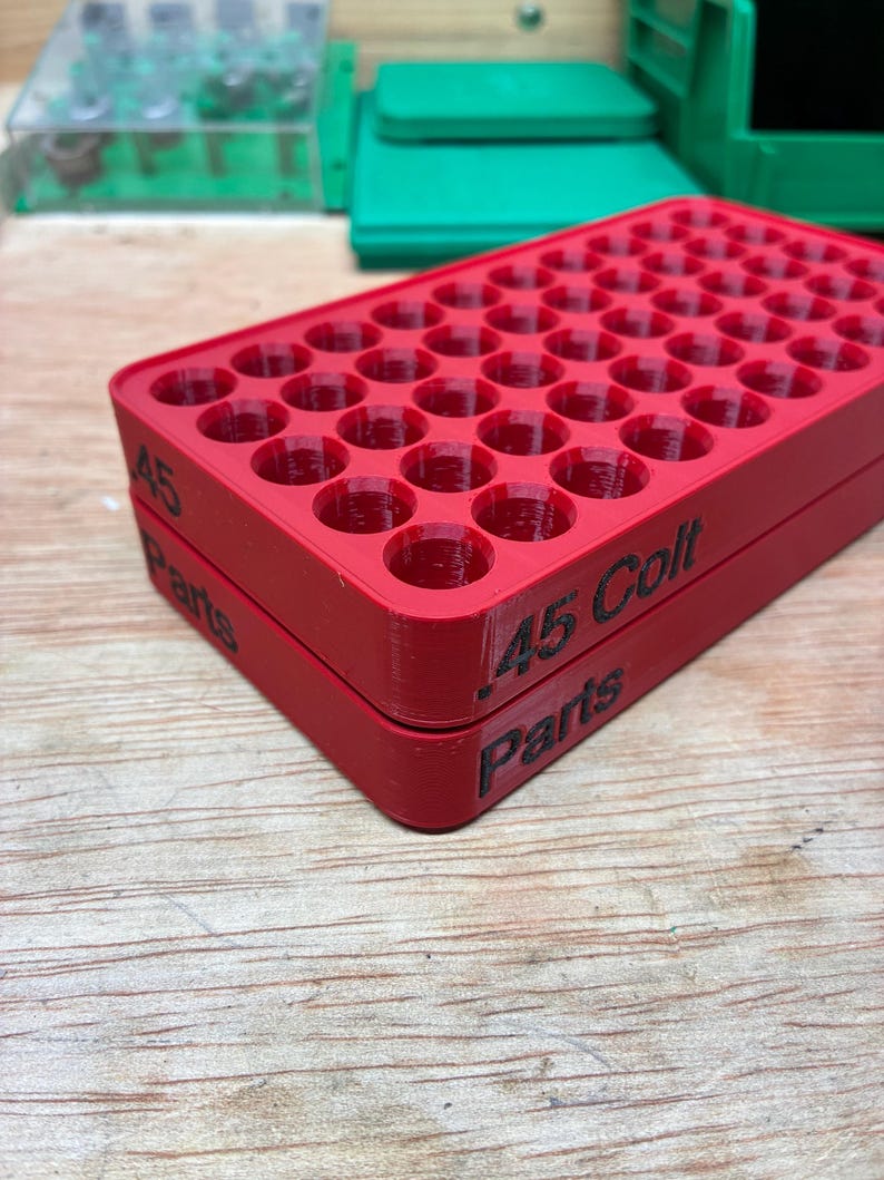 Pode incluir: Uma bandeja de armazenamento retangular de pl&aacute;stico vermelho com uma grade de orif&iacute;cios circulares para organizar muni&ccedil;&otilde;es. A lateral da bandeja apresenta texto impresso em preto que diz .45 Colt e Parts. Design estruturado e em camadas.