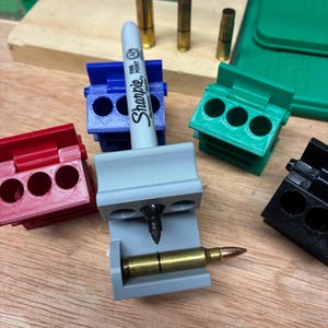 May include: A gray plastic cartridge case trimming jig holding a Sharpie fine point permanent marker and a brass rifle cartridge. The jig features a horizontal slot for the cartridge and a vertical hole for the marker. Additional red, blue, green, and black jigs are arranged nearby.