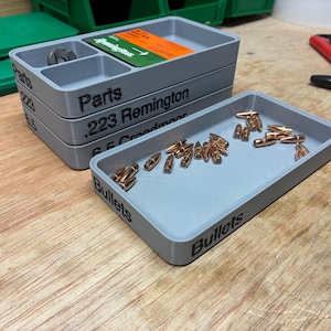 Pode incluir: Uma pilha de tr&ecirc;s bandejas de armazenamento retangulares de pl&aacute;stico cinza com etiquetas de texto preto lendo Parts, .223 Remington e 6.5 Creedmoor, ao lado de uma bandeja cinza individual rotulada Bullets contendo numerosos proj&eacute;teis de metal cor de cobre.