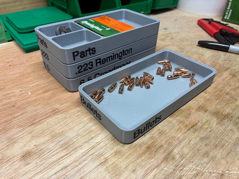 Pode incluir: Uma pilha de tr&ecirc;s bandejas de armazenamento retangulares de pl&aacute;stico cinza com etiquetas de texto preto lendo Parts, .223 Remington e 6.5 Creedmoor, ao lado de uma bandeja cinza individual rotulada Bullets contendo numerosos proj&eacute;teis de metal cor de cobre.
