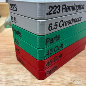 Pode incluir: Uma pilha de cinco bandejas retangulares de armazenamento de pl&aacute;stico nas cores cinza, verde e vermelho. Cada bandeja apresenta etiquetas de texto preto na lateral, incluindo .223 Remington, 6.5 Creedmoor, Parts, .45 Colt e Parts, indicando o conte&uacute;do.