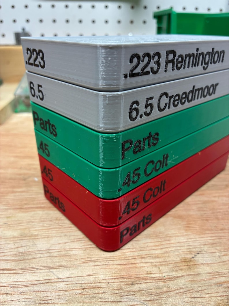 Pode incluir: Uma pilha de cinco bandejas retangulares de armazenamento de pl&aacute;stico nas cores cinza, verde e vermelho. Cada bandeja apresenta etiquetas de texto preto na lateral, incluindo .223 Remington, 6.5 Creedmoor, Parts, .45 Colt e Parts, indicando o conte&uacute;do.