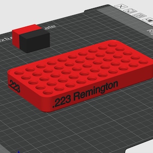 Pode incluir: Uma bandeja retangular de pl&aacute;stico vermelho para armazenamento de muni&ccedil;&atilde;o, com uma grade de orif&iacute;cios circulares para cartuchos. A lateral da bandeja tem impresso em preto o texto .223 Remington. Um pequeno bloco vermelho e preto est&aacute; posicionado ao lado.