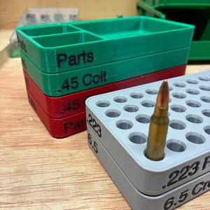 May include: A stack of rectangular plastic storage trays in green, red, and gray. The green tray is labeled Parts, the red tray is labeled .45 Colt, and the gray tray is labeled .223 and 6.5 Creedmoor, containing a single brass-cased cartridge.