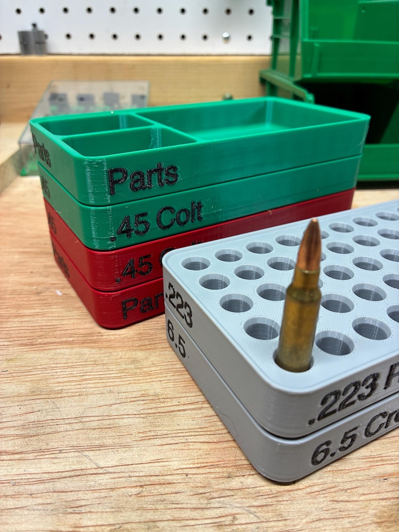 Pode incluir: Uma pilha de bandejas de armazenamento retangulares de pl&aacute;stico nas cores verde, vermelha e cinza. A bandeja verde est&aacute; marcada como Parts, a vermelha como .45 Colt e a cinza como .223 e 6.5 Creedmoor, contendo um cartucho de lat&atilde;o.