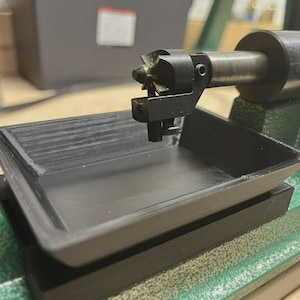 May include: A black tray sits on a green and black machine. A metal tool with a complex head is positioned above the tray. The machine appears to be used for precision work or manufacturing.