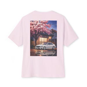 Könnte beinhalten: Hellrosa T-Shirt mit einem Aufdruck eines weißen Autos, das vor einem Haus mit einem Kirschblütenbaum geparkt ist. Die Spiegelung des Autos ist im nassen Boden zu sehen. Der Himmel ist blau mit Wolken. Das Shirt ist aus weichem Material.