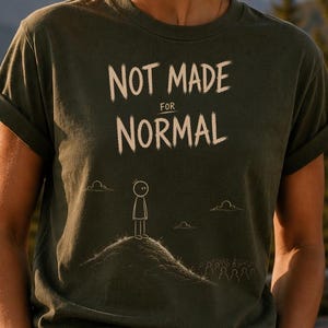 Op de afbeelding: Een olijfgroen T-shirt met korte mouwen en de witte tekst "NOT MADE FOR NORMAL" boven een eenvoudige lijntekening van een stokfiguurtje dat op een heuvel staat. Het T-shirt heeft een ronde hals en opgerolde mouwen.
