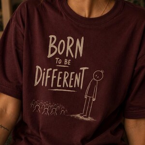 Op de afbeelding: Een donkerbordeauxrood T-shirt met korte mouwen en de witte tekst Born to be Different boven een afbeelding van een enkel stokfiguurtje dat apart staat van een grote menigte andere stokfiguurtjes, allemaal in handgetekende stijl op de borst.