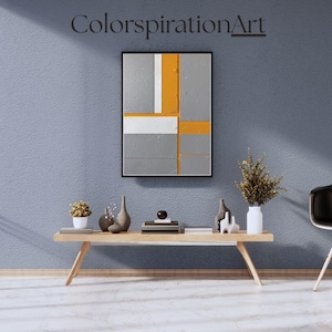 May include: A modern abstract painting with a black frame, featuring geometric shapes in gray, white, and orange. The artwork hangs on a textured blue wall above a wooden table with decorative items, including vases and a plant. The text "ColorspirationArt" is above the painting.