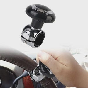 May include: A black and chrome steering wheel spinner. The spinner features a black, oval-shaped top with silver detailing and a chrome clamp. The image shows the spinner attached to a brown steering wheel.