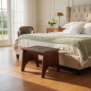 May include: A wooden step stool with a dark brown finish and a black top sits in front of a bed. The bedroom has hardwood floors, a tufted headboard, and a green knitted blanket on the bed. A gold lamp and a vase of white flowers are on a nightstand.