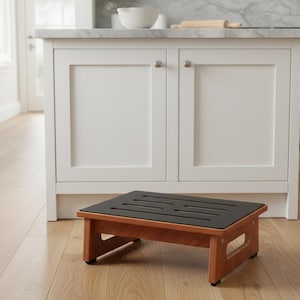 May include: A wooden step stool with a dark brown top and a light brown frame. The top has several evenly spaced slots. The stool is placed in front of a white kitchen island with a marble countertop. The floor is light-colored wood.