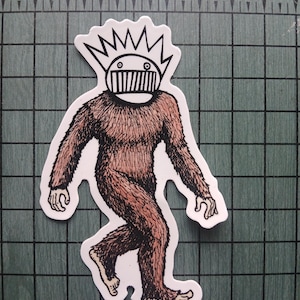 May include: A sticker featuring a cartoon illustration of a Bigfoot-like creature. The figure is brown and furry, with a white face and a spiky crown. The creature is walking, with its arms and legs outstretched.