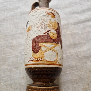 May include: A ceramic vase with a brown base and a cream-colored body. The vase features a painted scene of a person playing a harp. The handle and top are brown. The vase has a distressed, aged appearance.