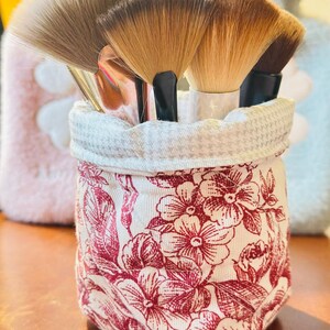 Könnte beinhalten: Eine Sammlung von Make-up-Pinseln mit hellbraunen Borsten und schwarzen Griffen wird in einem Behälter mit floralem Muster präsentiert. Der Behälter ist weiß mit einem roten Blumenmuster und einem Hahnentritt-Besatz.