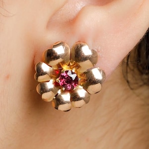 May include: A gold-colored flower-shaped stud earring with a small, round, pink gemstone in the center. The earring is worn on a person's ear, with the focus on the jewelry's design and color.