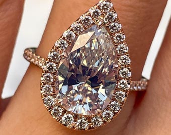 May include: Rose gold engagement ring featuring a pear-shaped diamond with a halo of smaller diamonds. The ring's band is also adorned with small diamonds, creating a sparkling effect. The ring is shown on a person's finger.