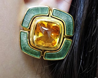 May include: A close-up of a gold-toned earring with a square, amber-colored gemstone at its center. The earring features a green enamel border with a geometric design. The earring is set against a person's ear and dark hair.