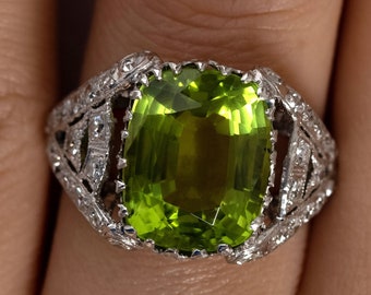 May include: A vintage-style ring with a large, green peridot gemstone set in a filigree design. The ring is made of white metal and has small diamonds surrounding the peridot.