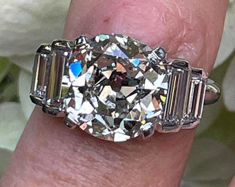 Puede incluir: Un anillo de tono plateado con una gran piedra central redonda facetada, flanqueada por dos piedras laterales rectangulares de corte escalonado. La banda de metal es pulida y reflectante, mostrando un diseño clásico de tres piedras.