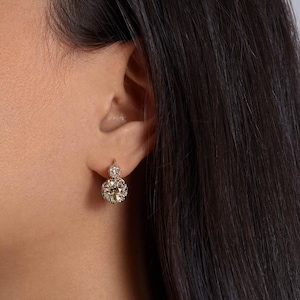 May include: A close-up of a person's ear wearing a pair of diamond drop earrings. The earrings feature a large, round, sparkling diamond at the bottom and a smaller diamond at the top, set in a gold or platinum setting.